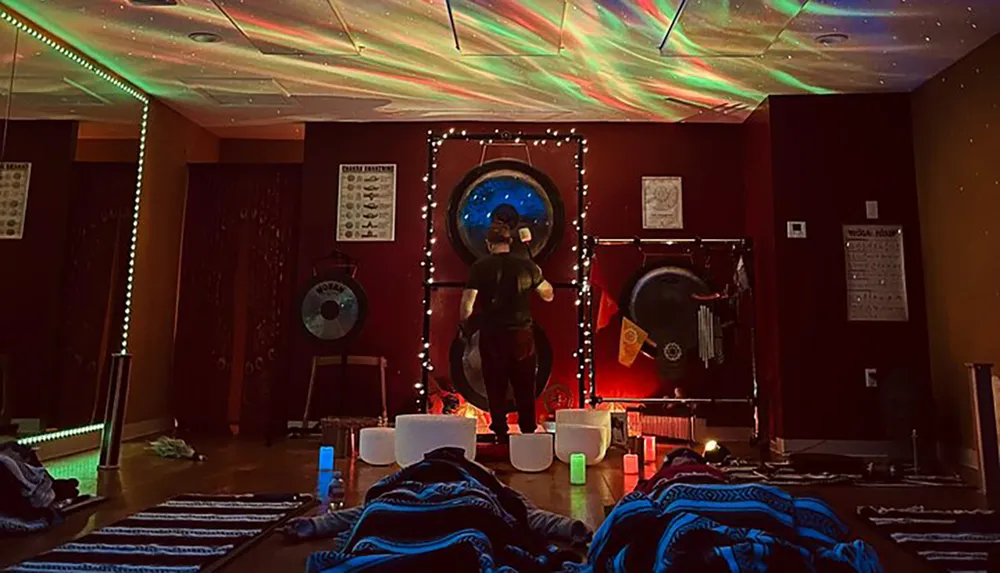 The image shows a dimly lit room with a person in the center playing a gong surrounded by lights and what appear to be meditation or relaxation aids under a ceiling with colorful light projections