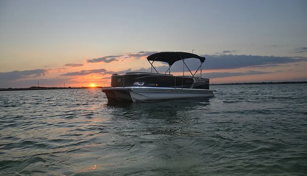 A pontoon boat floats on calm waters against the backdrop of a beautiful sunset