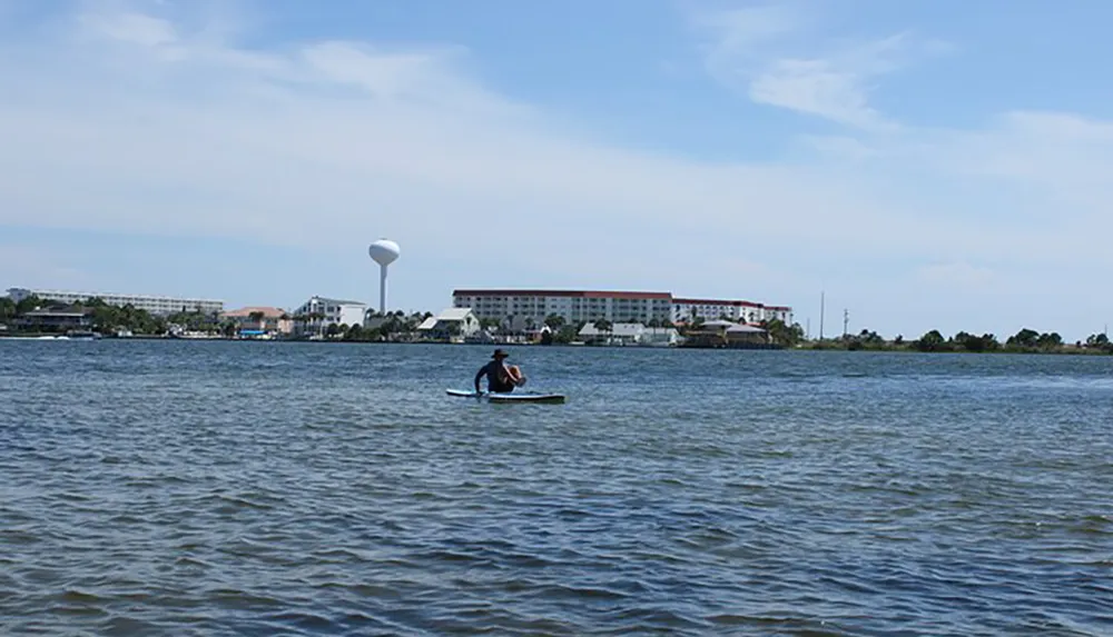 A person is paddleboarding on a calm body of water with buildings and a water tower in the background