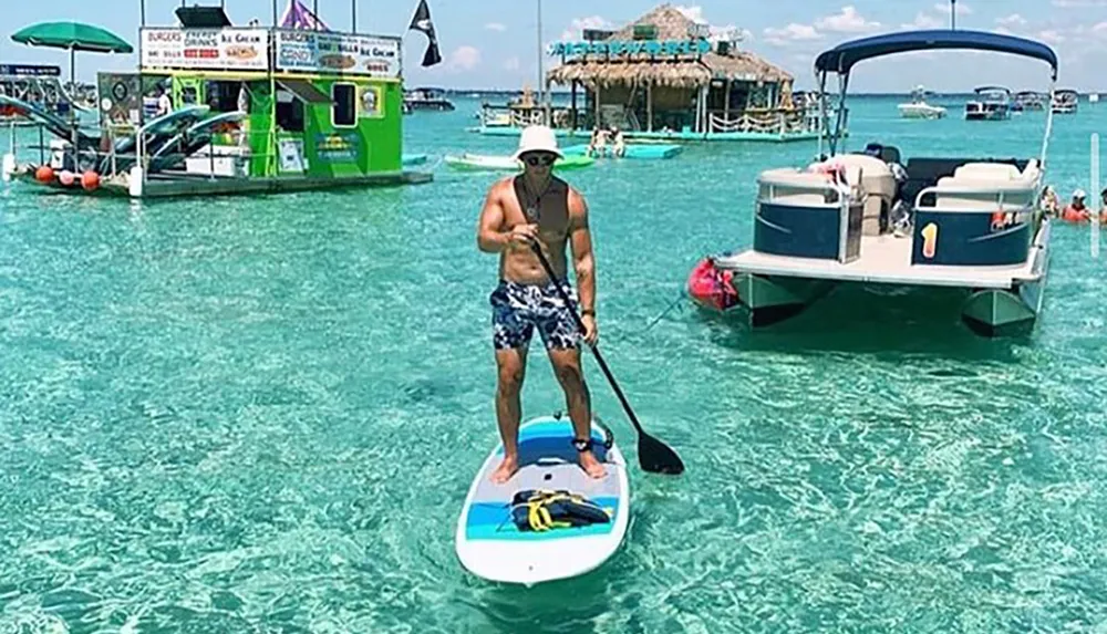 A person is stand-up paddleboarding on clear turquoise waters near a floating bar and other boats