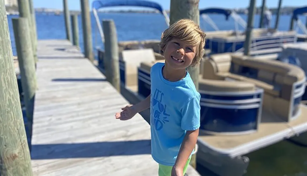 A smiling child stands on a sunny dock with boats moored in the background