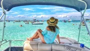 A person sits on the stern of a boat, gazing at a vibrant beach scene with clear turquoise water and numerous boats and people enjoying water activities.