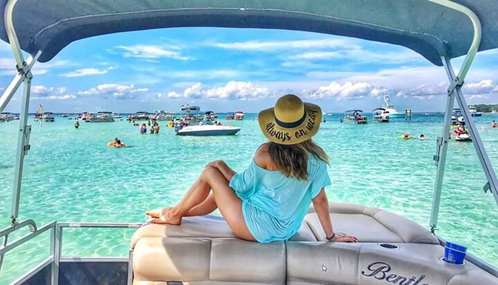 A person sits on the stern of a boat gazing at a vibrant beach scene with clear turquoise water and numerous boats and people enjoying water activities