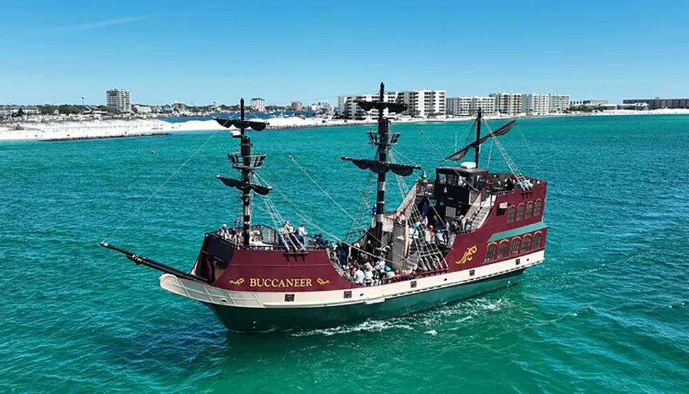 A modern replica of a pirate ship labeled BUCCANEER filled with people is sailing near a coastline with clear turquoise waters and a beachfront lined with buildings