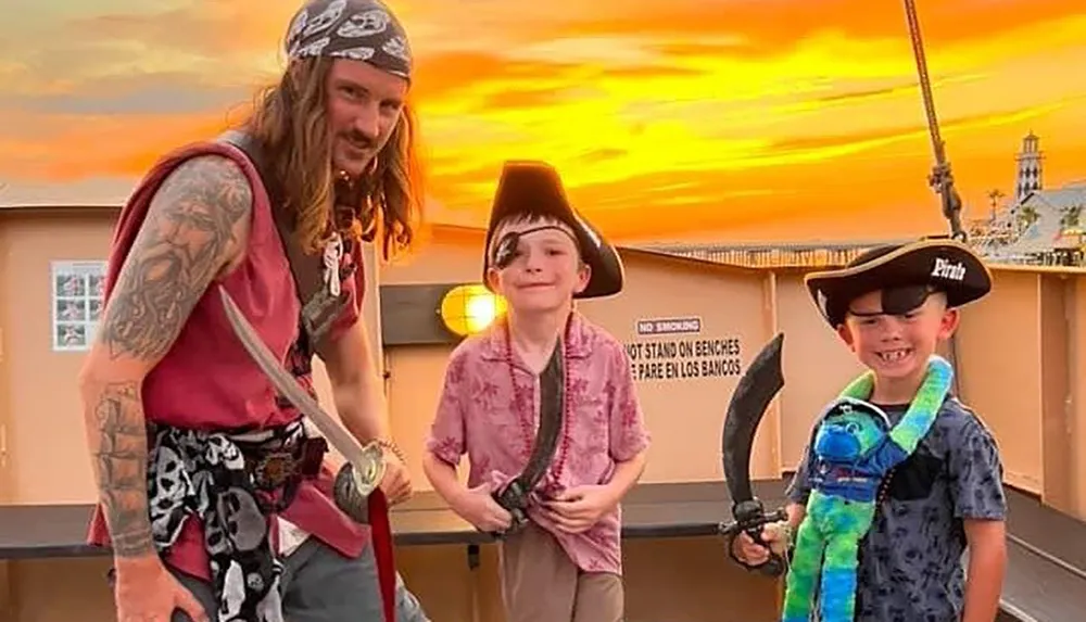 An adult and two children are dressed as pirates smiling and holding toy swords with a vivid sunset sky in the background