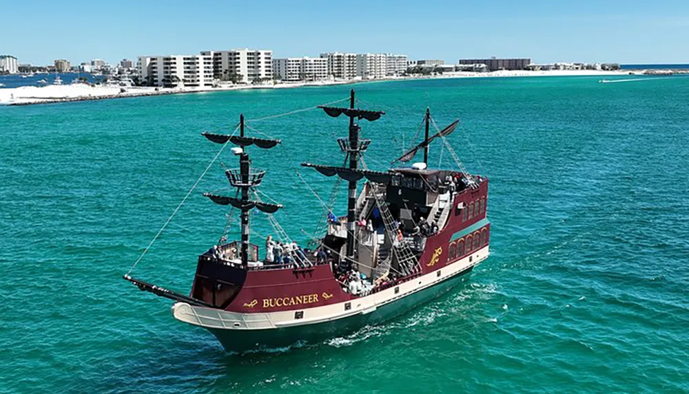 A pirate-themed tourist boat sails on turquoise waters near a coastline dotted with buildings