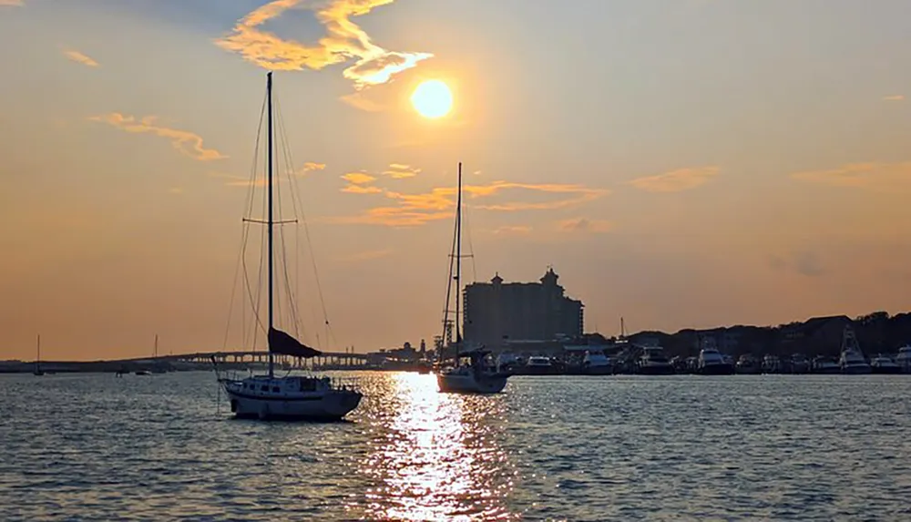 The image shows a serene sunset over a harbor with sailboats reflecting golden light on the water and silhouettes of waterfront buildings against a dusky sky