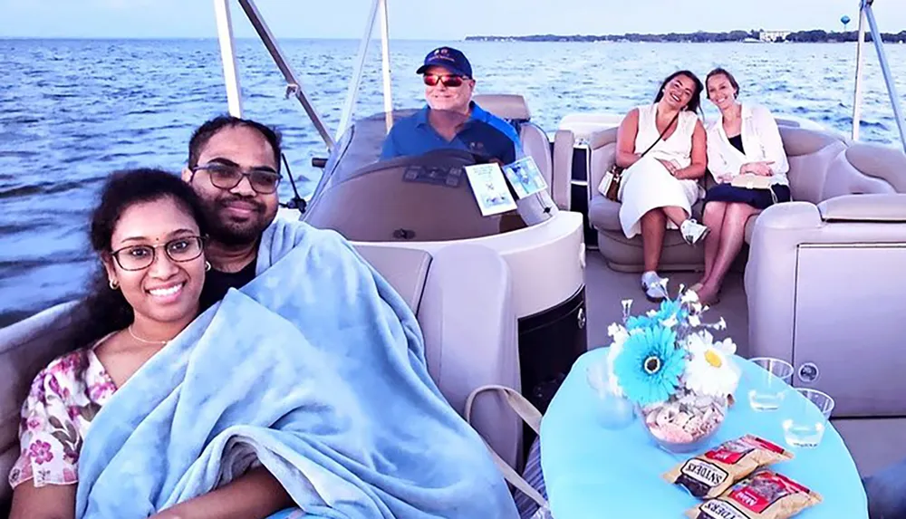 A group of people appears to be enjoying a boat trip with a couple wrapped in a blanket at the forefront a man steering the boat and two women seated at the back smiling