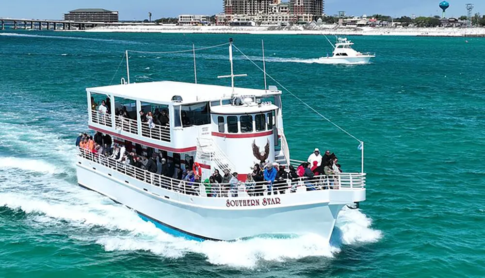 A double-deck tour boat named Southern Star filled with passengers is cruising in vibrant blue waters close to the shore with buildings in the background and another smaller boat speeding by in the distance