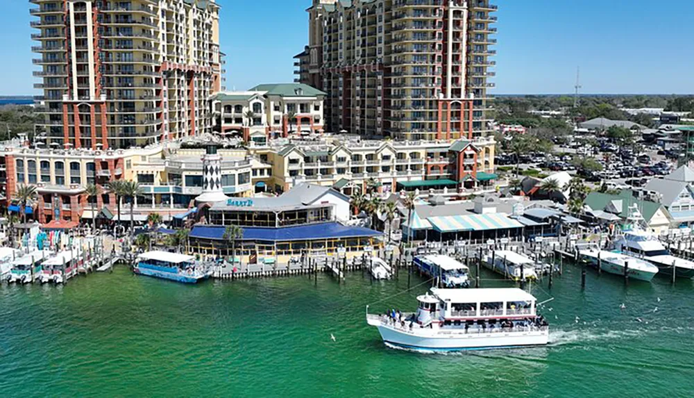 The image depicts a vibrant waterfront scene with colorful buildings a marina with several boats docked and a large tour boat cruising in the foreground set against a backdrop of high-rise condos under clear blue skies