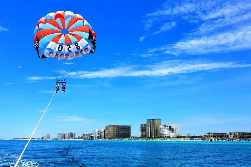 Two people are parasailing over a blue ocean in front of a skyline of coastal buildings under a clear sky