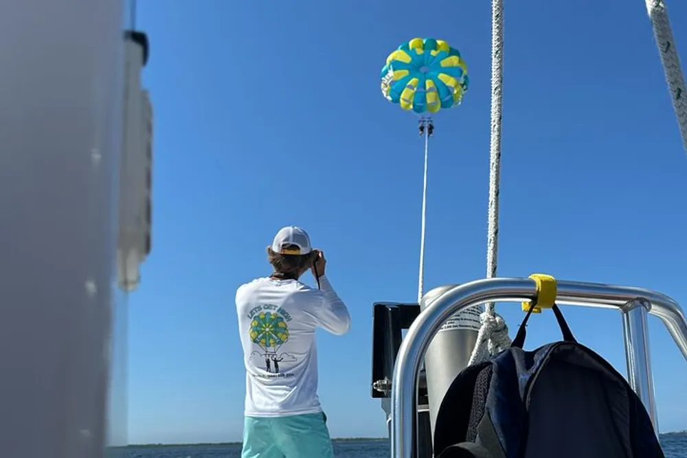 A person wearing a white cap and t-shirt is standing on a boat taking a picture with a camera next to a colorful spinning wind indicator under a clear blue sky