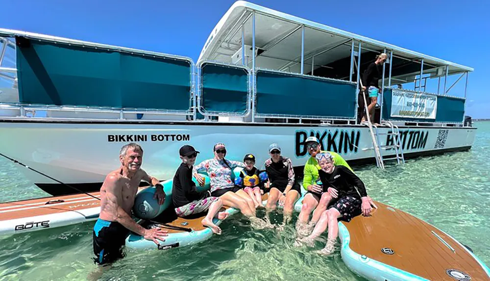 A group of people is enjoying the clear shallow waters next to a boat named BIKKINI BOTTOM with some sitting on a paddleboard and others standing or preparing to swim