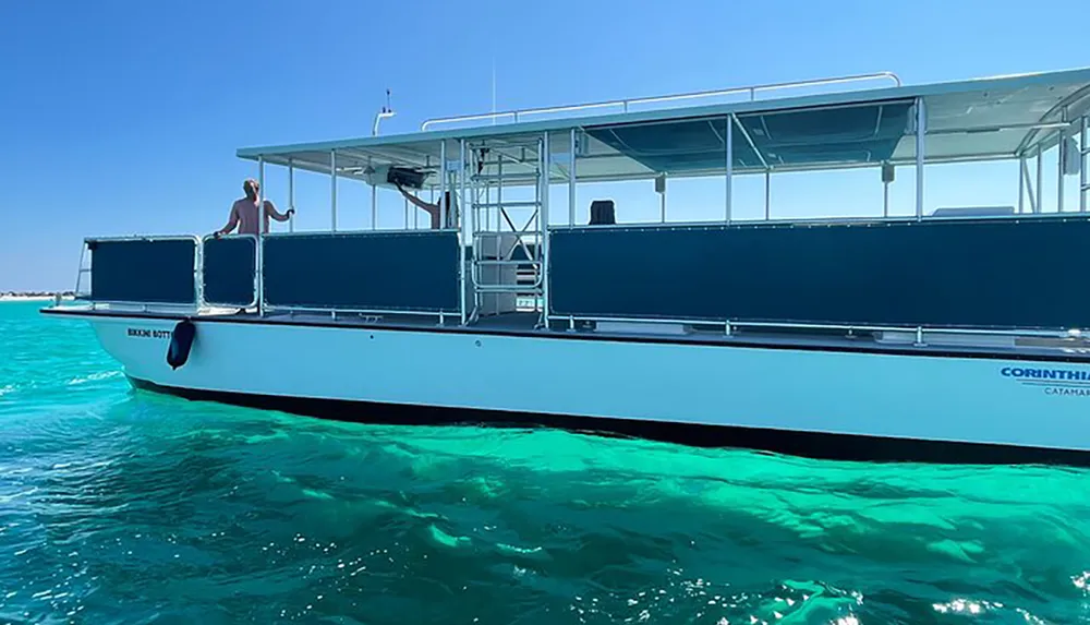 A person stands on a modern double-hulled catamaran boat as it floats on clear turquoise water under a bright blue sky