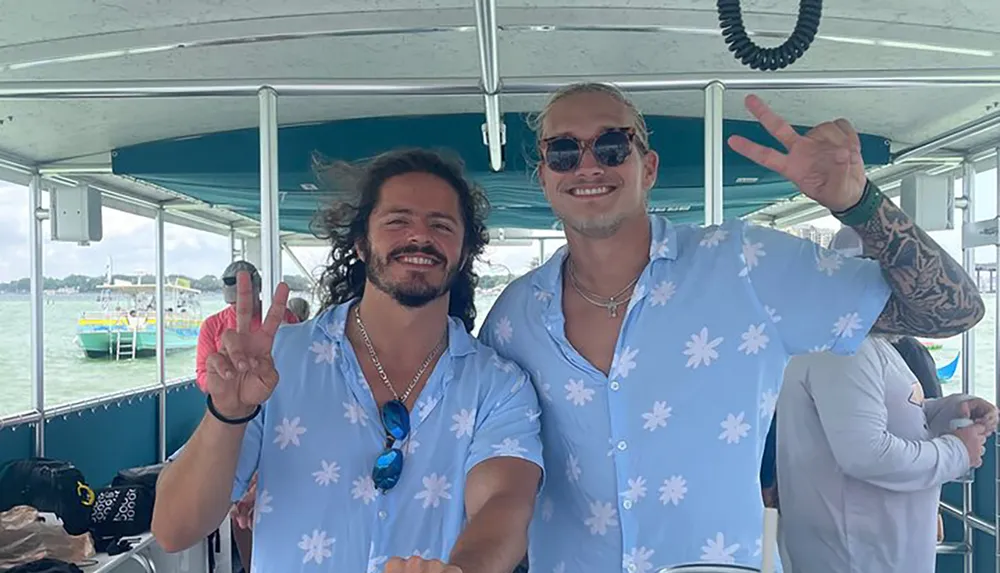 Two smiling men in matching floral shirts are making peace signs aboard a boat with a water backdrop