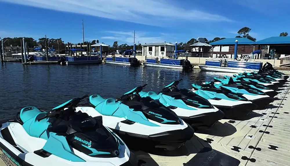 A row of turquoise and white jet skis are lined up on a dock with boats and waterfront buildings in the background under a clear blue sky