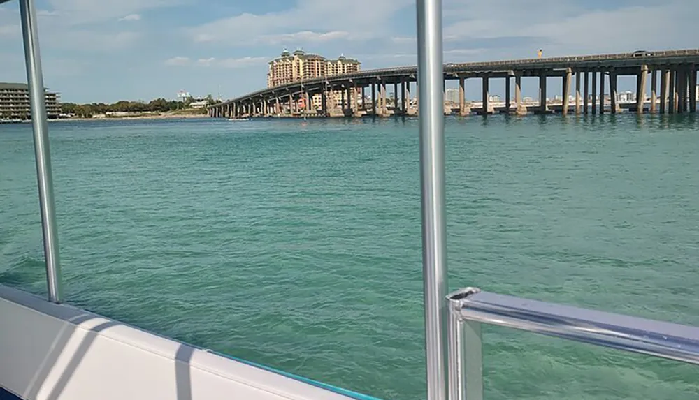 The image shows a tranquil turquoise waterfront with a bridge in the background viewed from the side of a boat framed by its metal railings