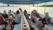 A group of people are enjoying a boat tour during sunset, with some sitting on benches and others standing, surrounded by calm waters.