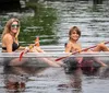 A smiling woman and a young boy are sharing a clear kayak both holding paddles on a calm water body