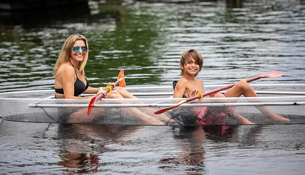 A smiling woman and a young boy are sharing a clear kayak both holding paddles on a calm water body