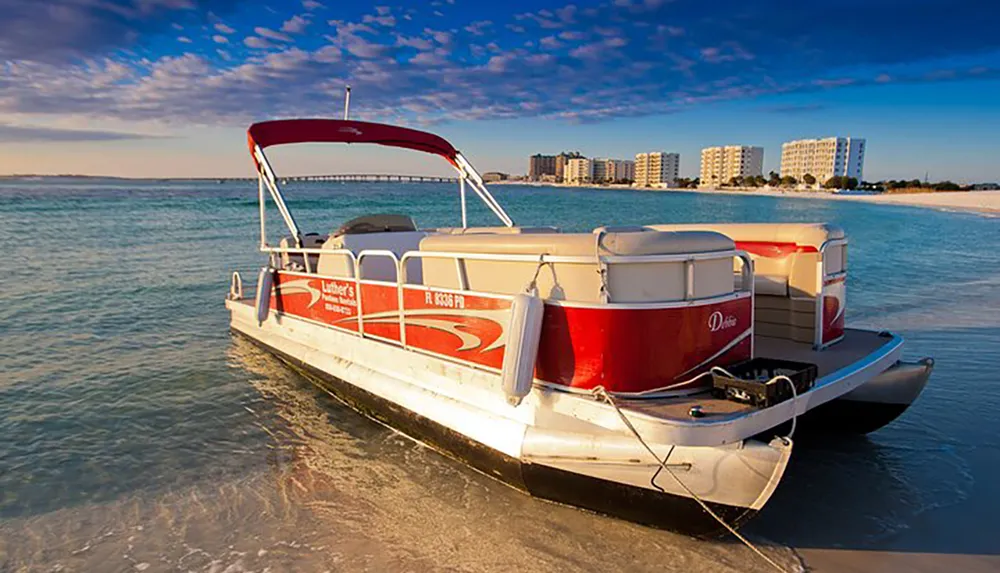 A red and white pontoon boat is beached on a clear sandy shore with a backdrop of a sunny sky calm water and distant apartment buildings