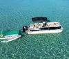 The image shows a lively water scene with people enjoying various activities on boats a floating mat and a paddleboard in clear turquoise waters