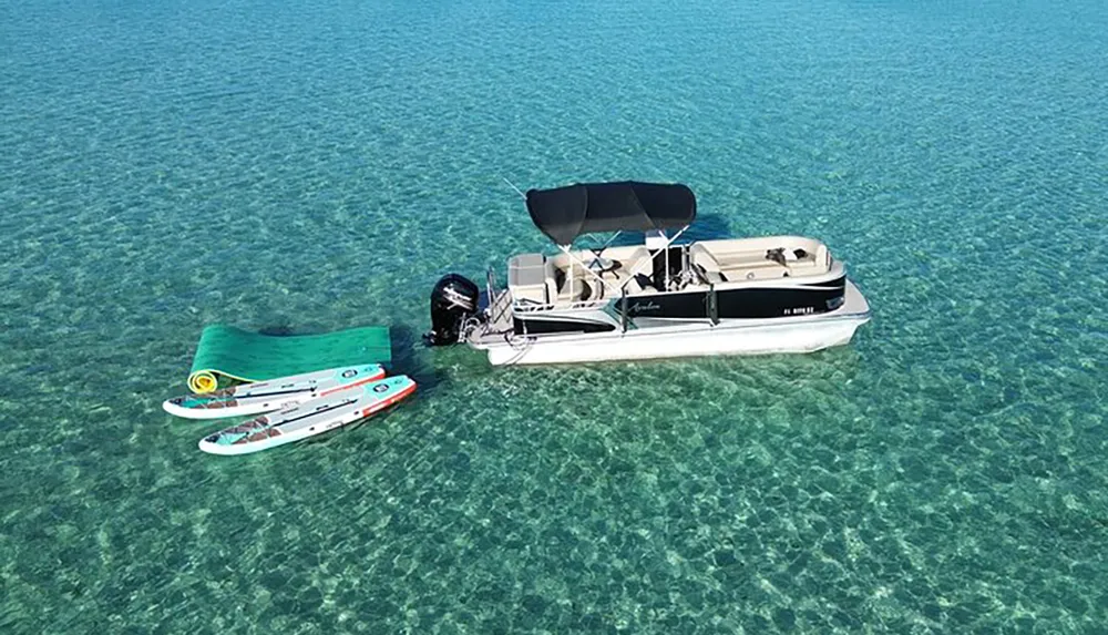 A pontoon boat is anchored in crystal-clear shallow water near two paddleboards suggesting a leisurely day on the water