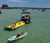 The image shows a lively water scene with people enjoying various activities on boats a floating mat and a paddleboard in clear turquoise waters