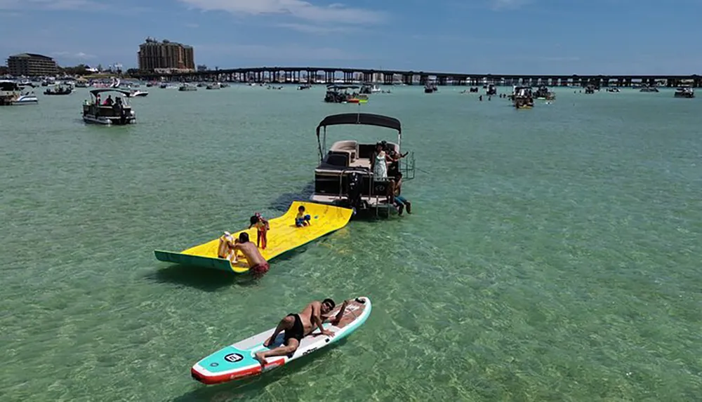 The image shows a lively water scene with people enjoying various activities on boats a floating mat and a paddleboard in clear turquoise waters