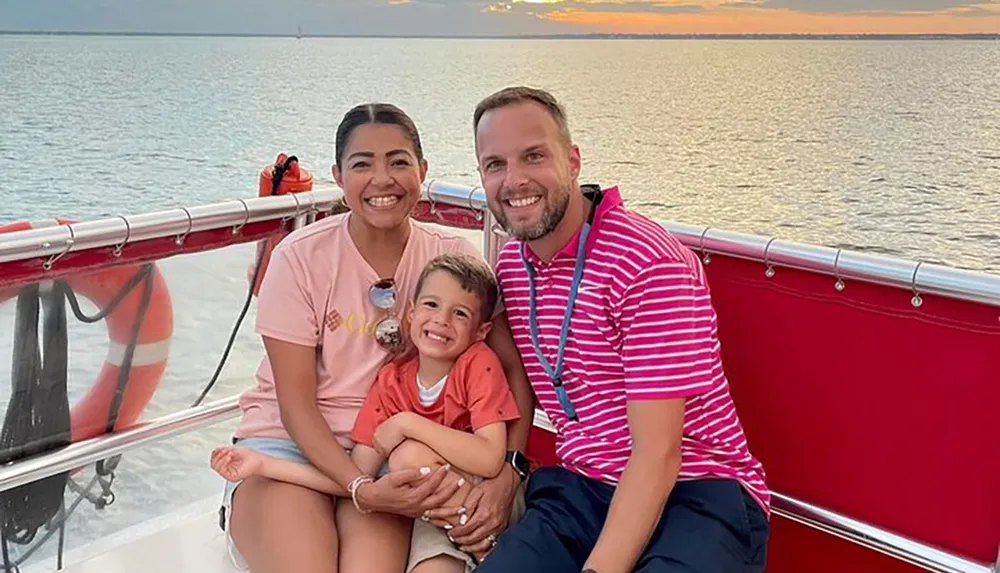 A happy family of three is smiling for a photo aboard a boat with a scenic sunset over the water in the background