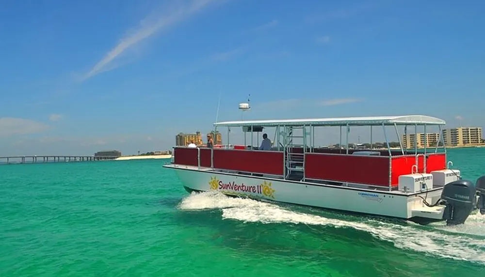 A pontoon boat named SunVenture II is cruising on bright turquoise waters near a sandy coastline with a pier and buildings in the background under a blue sky