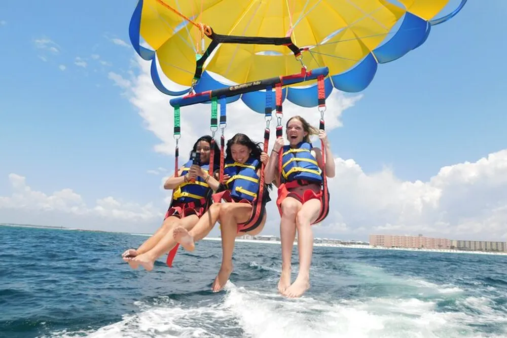 Three individuals are enjoying parasailing above the ocean with smiles on their faces suggesting they are having a great time