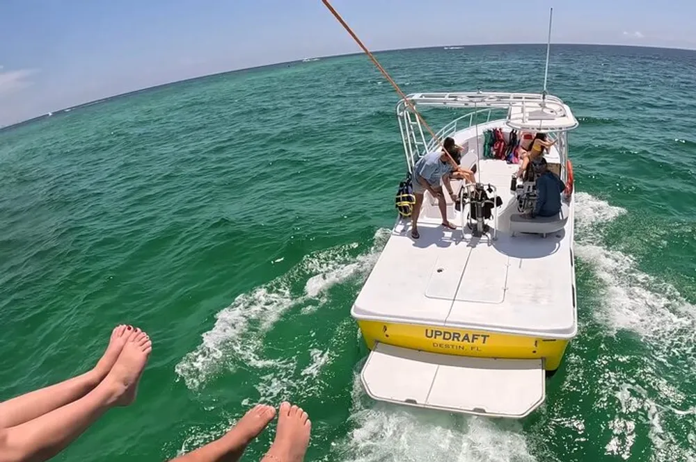 A group of people enjoys a sunny day on the deck of a boat cruising through greenish-blue waters with a pair of legs visible in the foreground suggesting someone is reclining and taking the photo