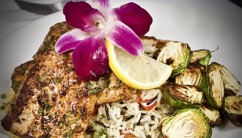 A plate of grilled fish garnished with a lemon wedge and an orchid accompanied by wild rice and roasted Brussels sprouts