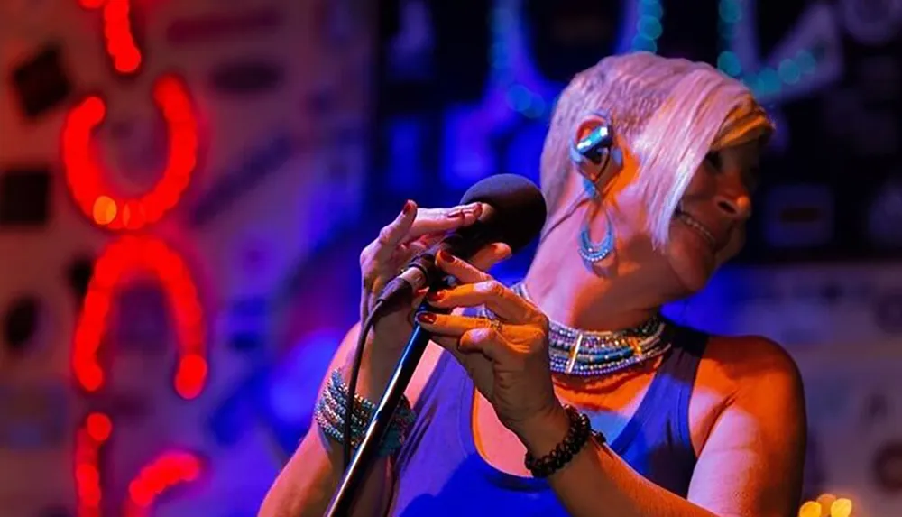 A singer with short blonde hair wearing jewelry holds a microphone and performs with emotion under colorful stage lighting