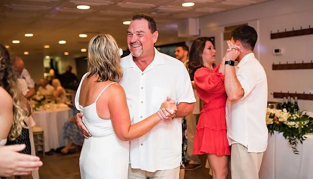 A couple in white attire are smiling at each other while dancing surrounded by other dancing guests at what appears to be a festive occasion possibly a wedding reception