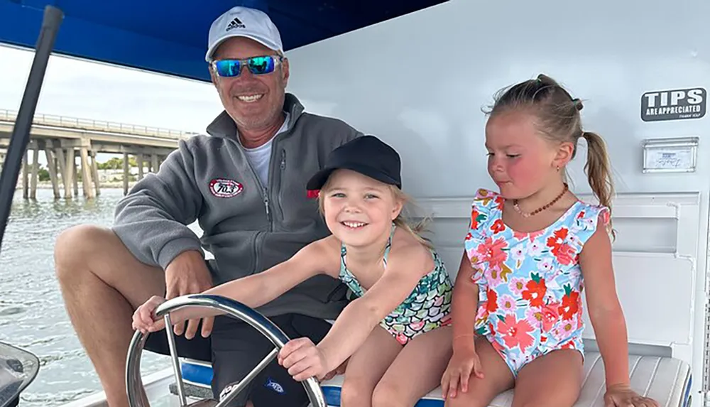 A man and two young girls are smiling and enjoying time on a boat with a bridge in the backdrop