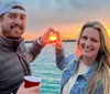 A man and a woman are creating a heart shape with their hands framing the setting sun over the water behind them