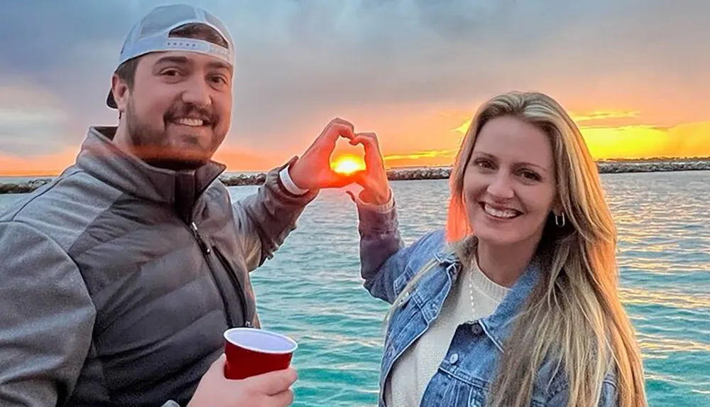 A man and a woman are creating a heart shape with their hands framing the setting sun over the water behind them