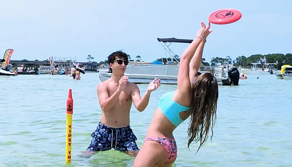 Two people are playing with a flying disc by the water with boats and other beachgoers visible in the background