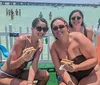 Three people are smiling for the camera while holding hot dogs sitting on a dock over turquoise water with other people wading and chatting in the background