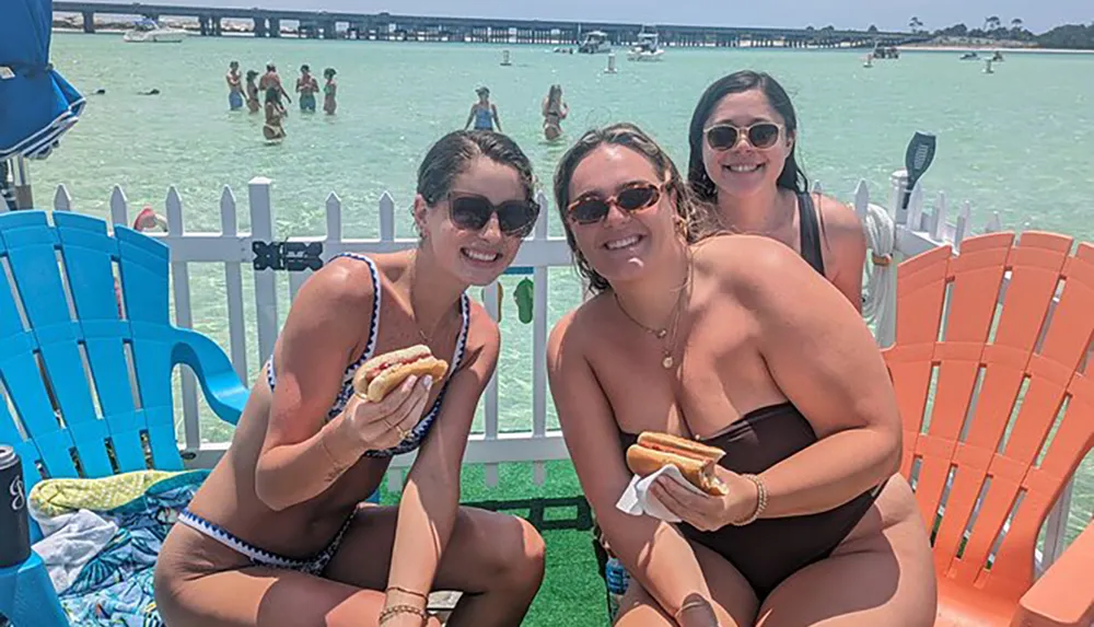 Three people are smiling for the camera while holding hot dogs sitting on a dock over turquoise water with other people wading and chatting in the background