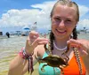 A smiling young woman is holding up a crab at a sunny waterfront setting with boats in the background