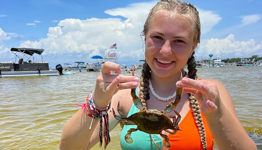 A smiling young woman is holding up a crab at a sunny waterfront setting with boats in the background