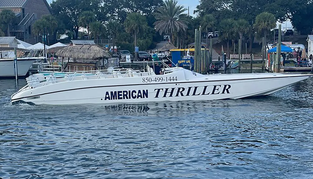 A white speedboat named AMERICAN THRILLER is docked on calm waters near a shore with palm trees and various structures