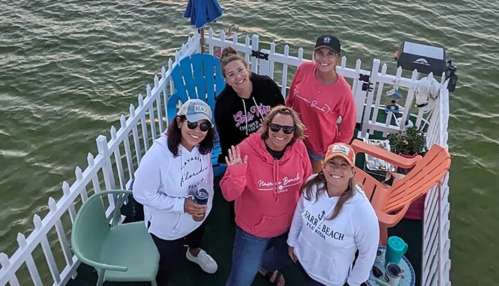 A group of five smiling women are posing together on a small floating platform with a white picket fence situated on a body of water