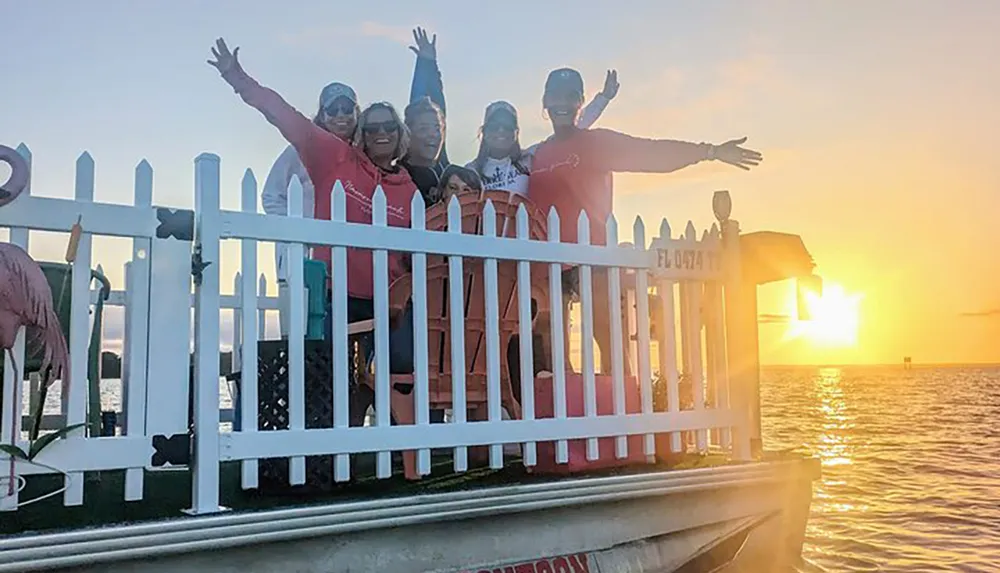 A group of smiling people is posing behind a white picket fence on a dock at sunset