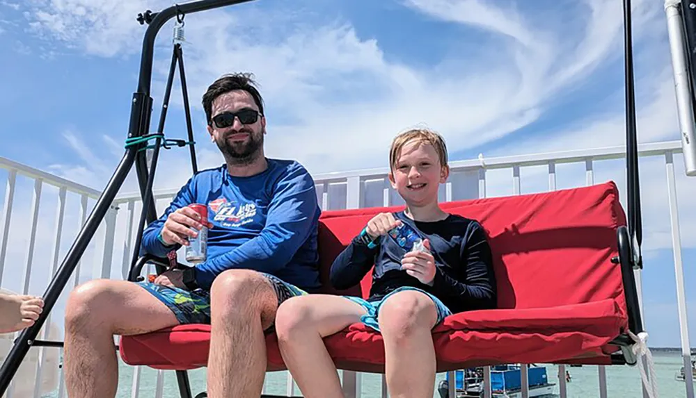 An adult and a child are sitting on a red swing by the sea both smiling and holding drinks on a sunny day