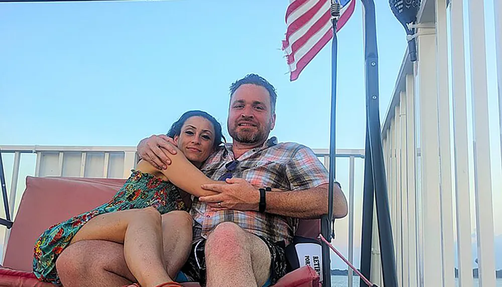 A couple is sitting close together on a balcony with an American flag in the background enjoying the outdoors