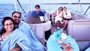 A group of people are enjoying a boat ride, with a couple snuggled under a blanket in the foreground and two women sitting with snacks at a table, while a man pilots the boat in the background.